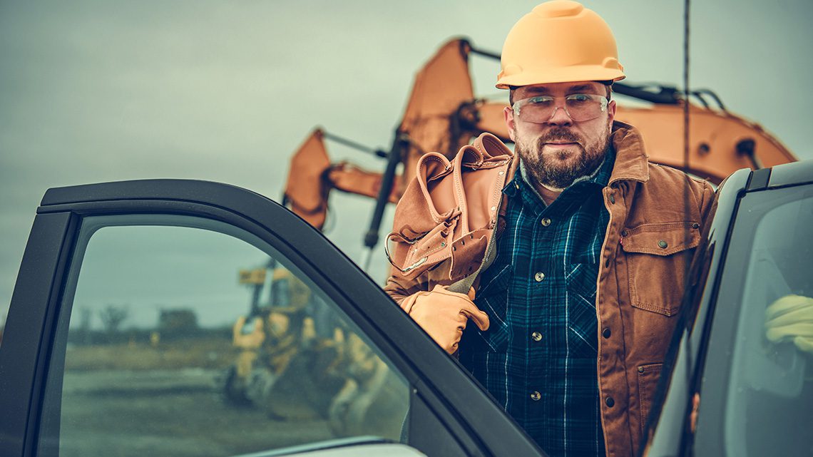 Man getting into a pickup truck on the job site with a yellow hardhat and tool belt